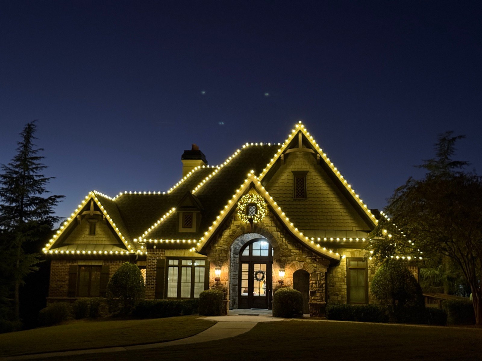 A stone and shingle Atlanta home at dusk, every gable and eave of its multi-plane roofline traced in warm white bulbs, a lit wreath in the front gable
