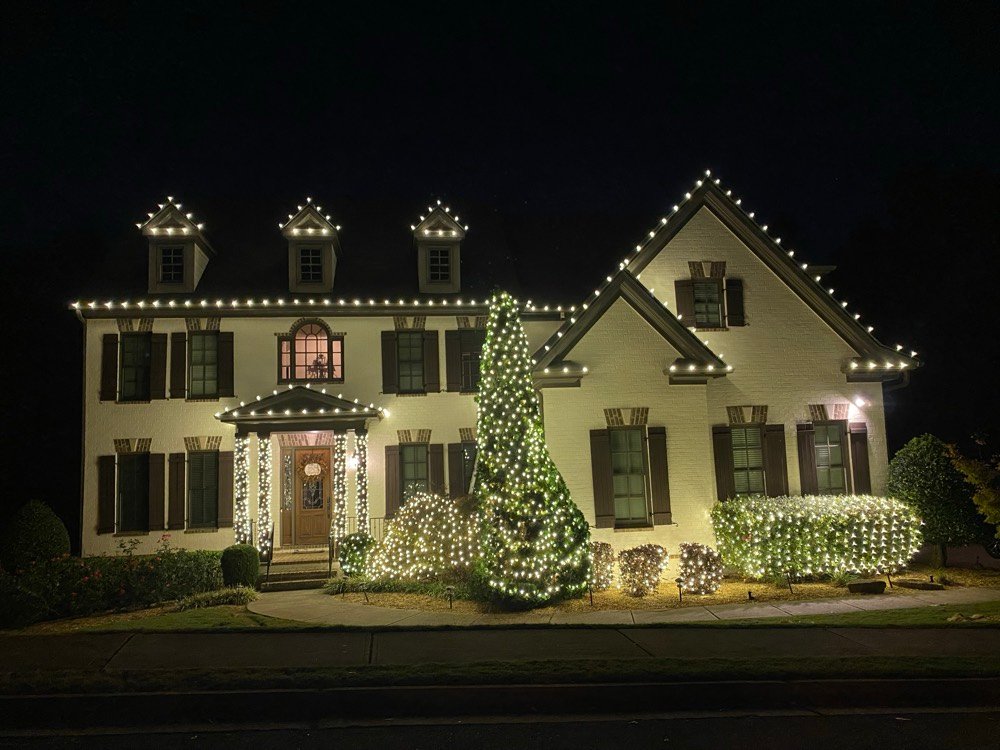 A white colonial at night — lit dormers and rooflines, garland-wrapped entry, a fully lit evergreen and netted shrub beds