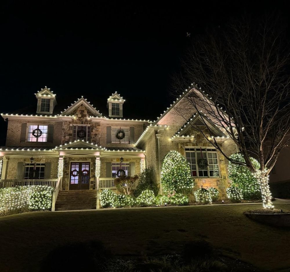 A metro Atlanta stone estate lit by Happy Lights of Atlanta — every gable and dormer of the roofline traced in warm white, wreaths in the windows, shrubs netted in light